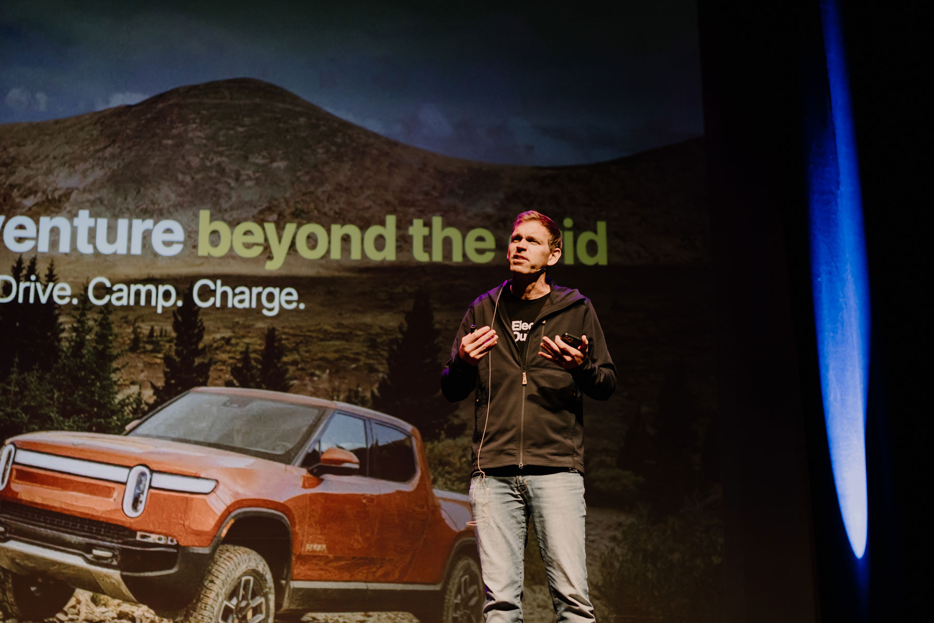 A man speaking on stage with a presentation on screen behind him