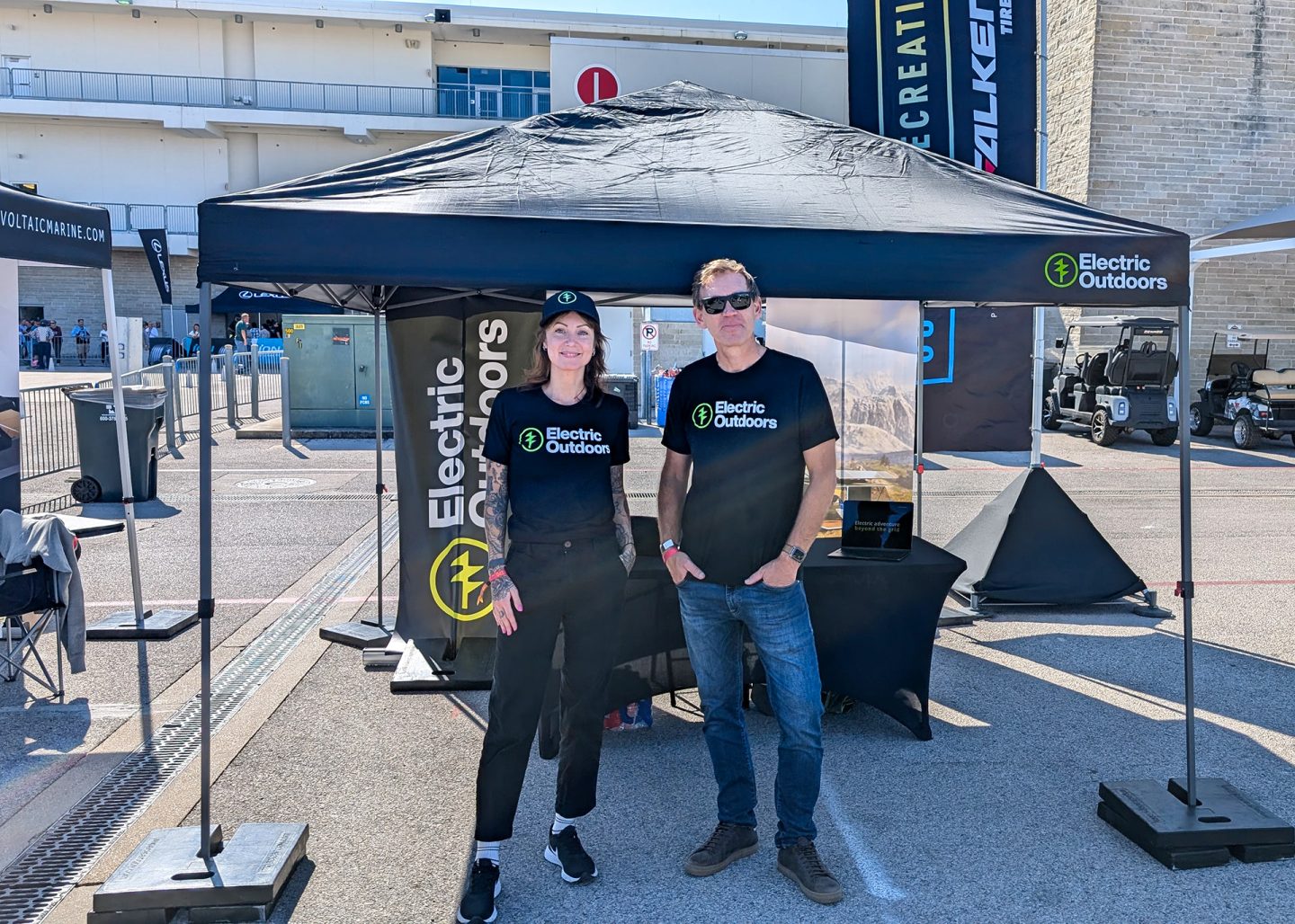 Two people wearing matching Electric Outdoors stand in front of a canopy tent at an event