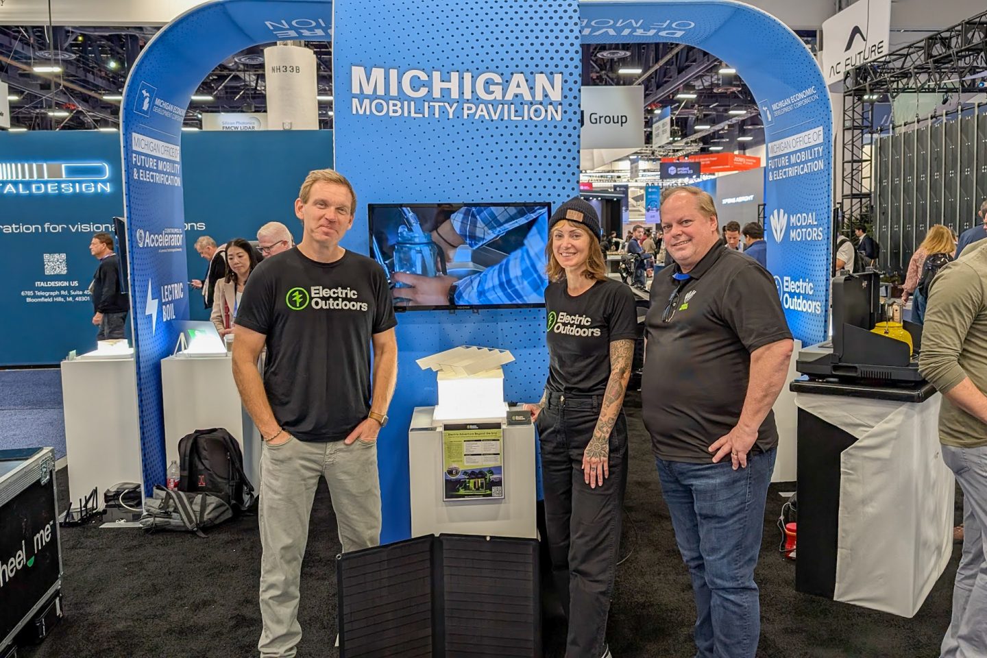 Two men and a woman stand in front of a trade show booth smiling at the camera