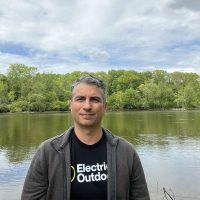 A man stands in front of a still lake with trees in the background