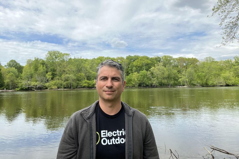 A man stands in front of a still lake with trees in the background